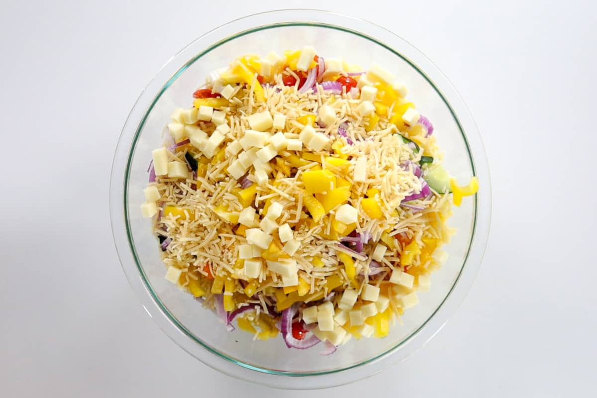 A glass bowl filled with gluten-free rotini pasta, tomatoes, cucumber, bell pepper, red onion, mozzarella cheese, and parmesan cheese.