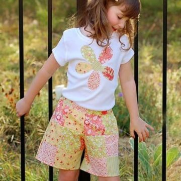 Young girl wearing a white shirt with a floral applique and colorful patchwork shorts, smiling and playing near a black metal fence in a sunny garden.
