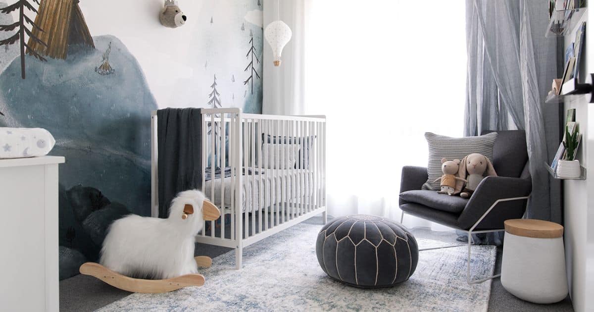 Woodland nursery with a white crib, mountain mural, gray reading chair, rocking sheep, plush toys, and a soft blue rug in a bright modern space.