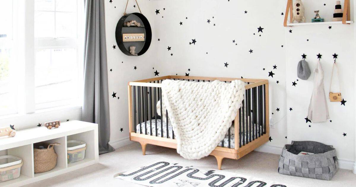 Modern monochrome nursery with a natural wood crib, chunky knit blanket, black star wall decals, gray curtains, and minimalist shelves.