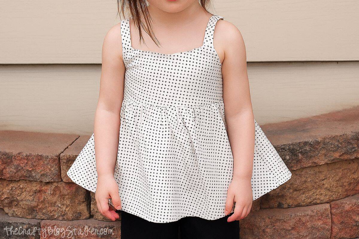 Toddler girl wearing a handmade white tank top with small black polka dots, standing in front of a brick and siding wall.