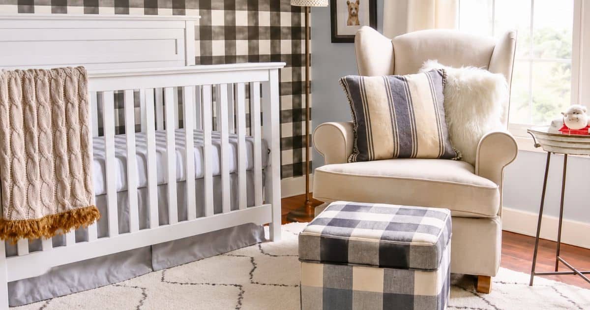 Neutral nursery with a white crib, cream wingback glider, buffalo plaid wallpaper, matching ottoman, and cozy knit blanket in a bright room.
