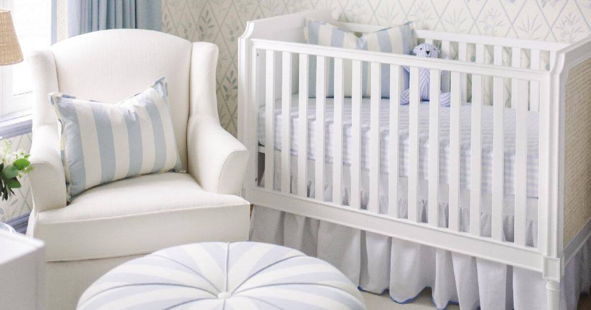 Blue and white nursery with a white crib, upholstered armchair, striped pillows, soft patterned wallpaper, and coordinating blue bedding.