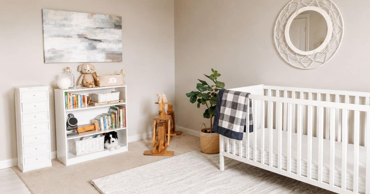 Neutral nursery with a white crib, white bookshelf, rocking horse, black and white plaid blanket, round wall mirror, and soft beige walls.