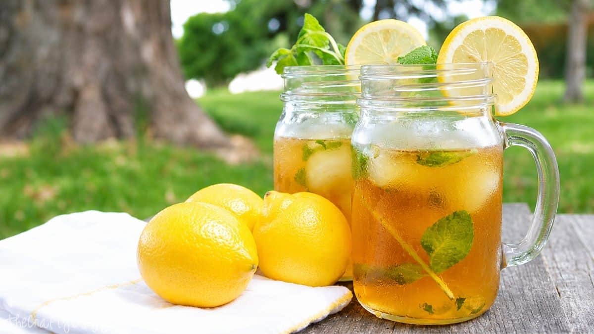 Two mason jars of iced tea with lemon slices and mint leaves, served with ice cubes, next to three fresh lemons on a towel outdoors.