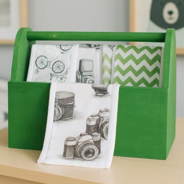 Green wooden caddy holding handmade burp cloths with printed fabric centers, including bicycles, cameras, and green chevron patterns.
