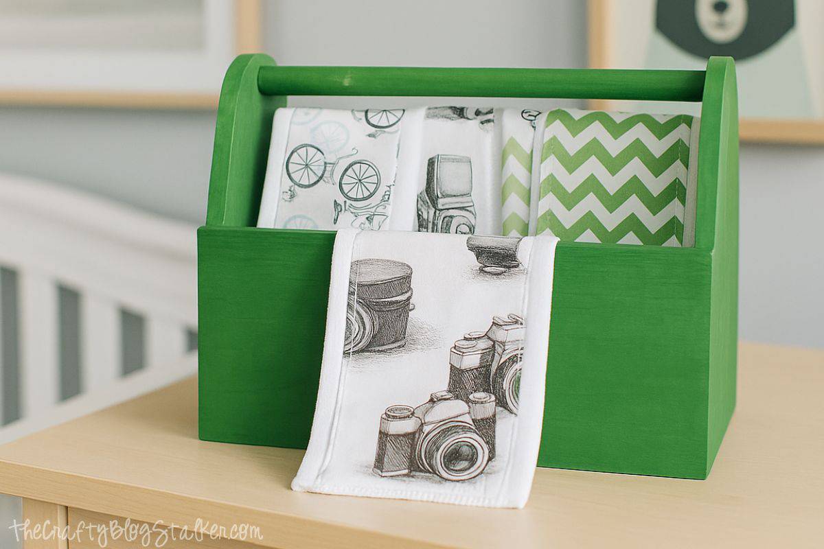 Green wooden caddy holding rolled burp cloths with bike, camera, and green chevron prints, displayed on a nursery dresser.