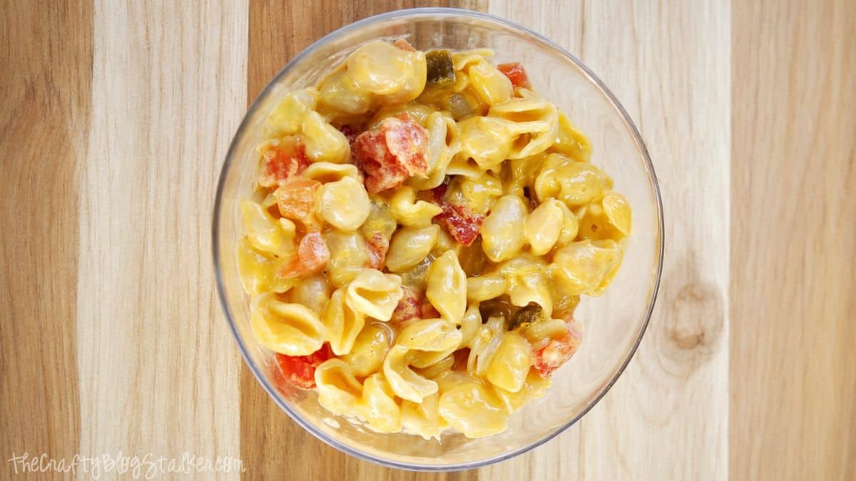 Top view of cheesy pasta shells mixed with diced tomatoes and peppers in a glass bowl on a wooden surface.