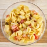 Close-up of cheesy pasta shells mixed with diced tomatoes and peppers in a glass bowl on a wooden surface.