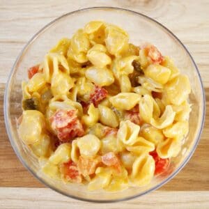 Close-up of cheesy pasta shells mixed with diced tomatoes and peppers in a glass bowl on a wooden surface.