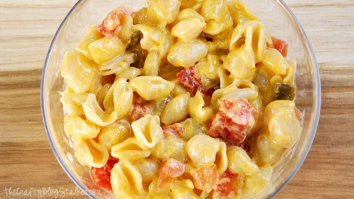Close-up of cheesy pasta shells mixed with diced tomatoes and peppers in a glass bowl on a wooden surface.