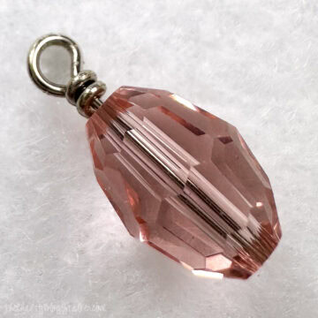 Close-up image of a faceted pink glass bead attached to a metal head pin with a loop at the top, resting on a soft white surface.