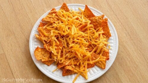 Plate of tortilla chips topped with shredded cheddar cheese, ready to be melted, on a wooden table.