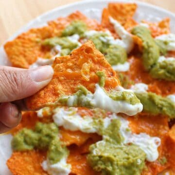 Hand holding a tortilla chip topped with melted cheese, guacamole, and sour cream over a plate of nachos.