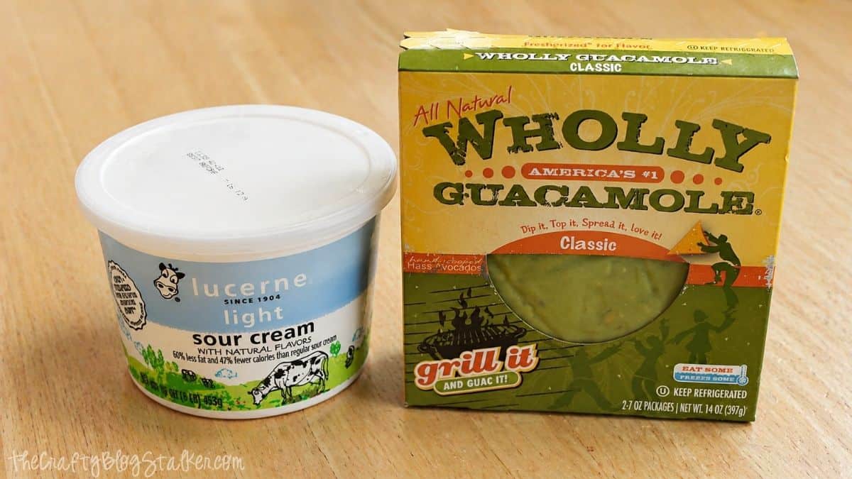 Container of sour cream and a box of Wholly Guacamole on a wooden table.