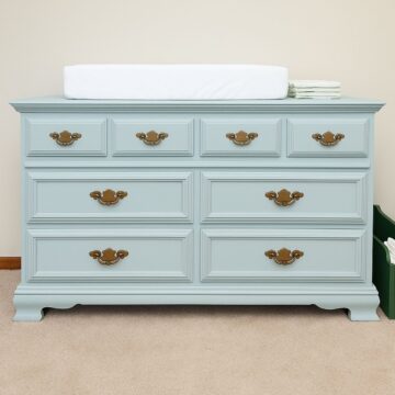 A vintage dresser painted in soft blue with brass handles, topped with a white changing pad and stacked diapers, sits on beige carpet against a cream wall.