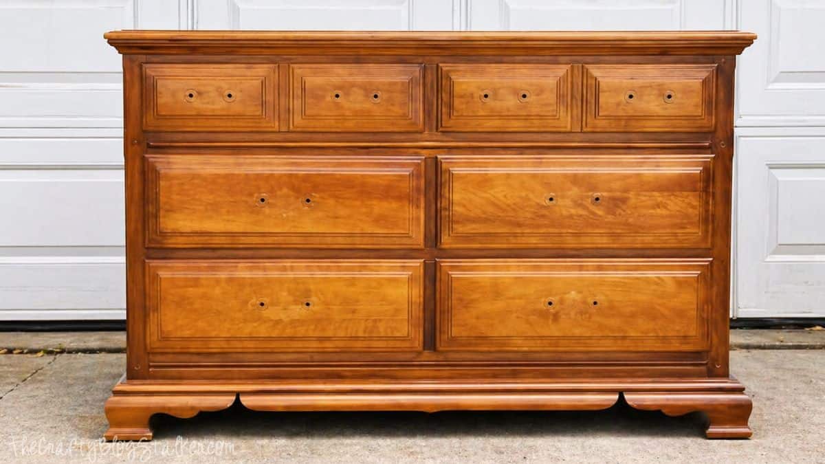 A wooden dresser with a glossy honey-brown finish and missing drawer hardware, photographed indoors against a white paneled wall.