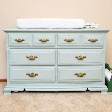 A vintage dresser painted in soft blue with brass handles, topped with a white changing pad and stacked diapers, sits on beige carpet against a cream wall.