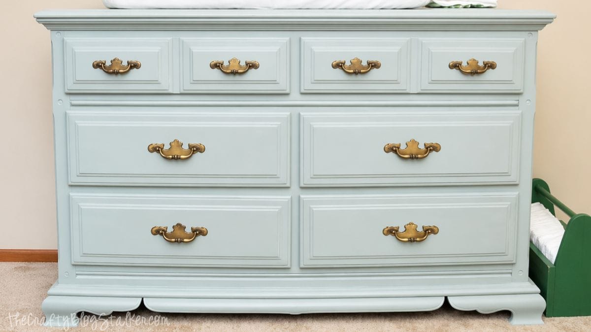 A refinished dresser painted in light blue with vintage brass handles, styled with a changing pad and neatly stacked diapers on top.