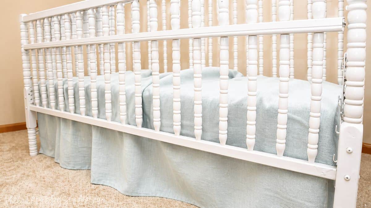 The Easiest DIY Crib Skirt Tutorial Ever Side view of a white crib with a light fabric skirt and mattress, set on carpet in a nursery room.