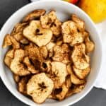 Close-up of homemade crispy apple chips sprinkled with cinnamon in a white bowl, with fresh apples in the background.