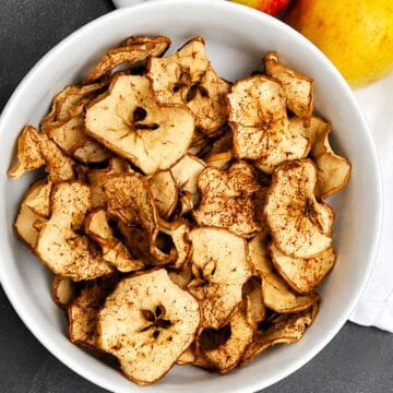 Close-up of homemade crispy apple chips sprinkled with cinnamon in a white bowl, with fresh apples in the background.