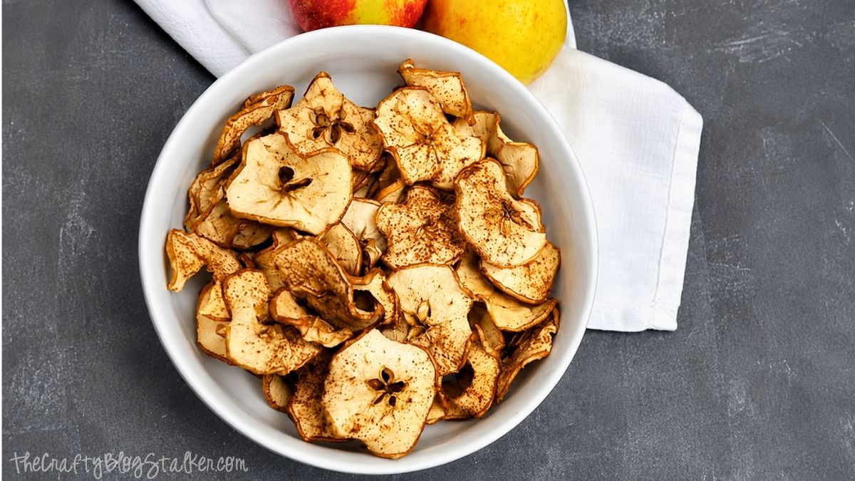 A yummy Apple Chips recipe great for this fall season! Enjoy! Homemade crispy apple chips sprinkled with cinnamon in a white bowl, with fresh apples on a dark countertop.