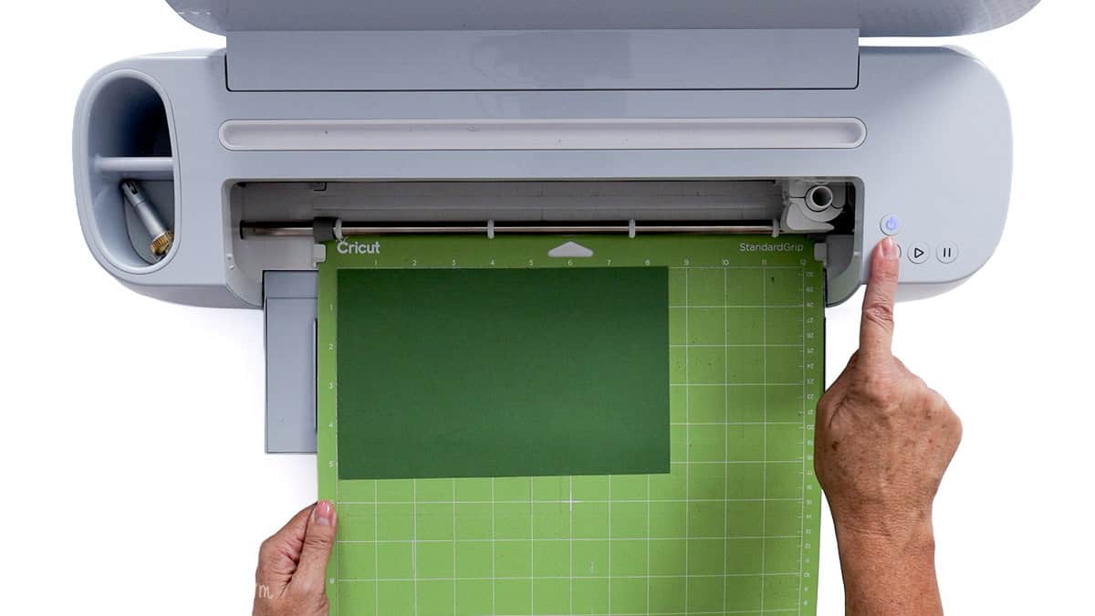 Loading a green Cricut mat with vinyl into a cutting machine to cut pieces for jack-o'-lantern candy jars.