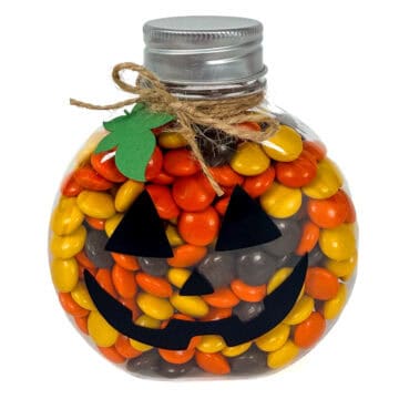 Clear pumpkin-shaped candy jar filled with orange, yellow, and brown candies, decorated with a black jack-o'-lantern face, twine, and green leaf.