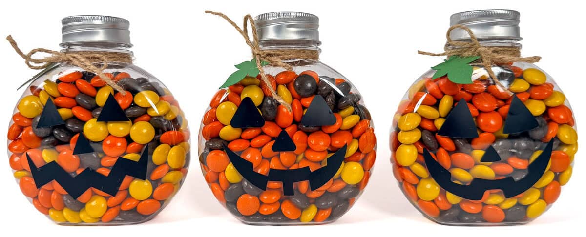 Three clear pumpkin-shaped jars filled with orange, yellow, and brown candy, decorated with black jack-o'-lantern faces and twine.