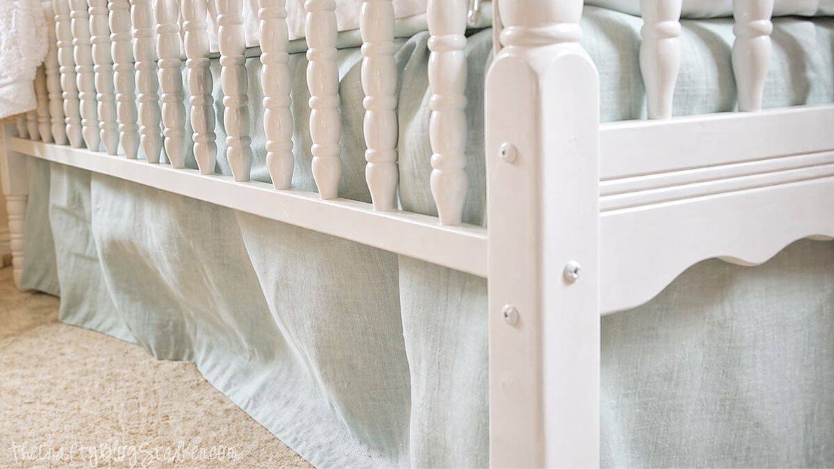 The Easiest DIY Crib Skirt Tutorial Ever Corner view of a white crib with a light fabric skirt and mattress, placed on carpet in a nursery room.