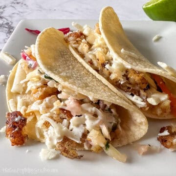 Two fish tacos on a white plate, filled with flaky fish, shredded cabbage, cheese, and creamy sauce, served with a lime wedge.