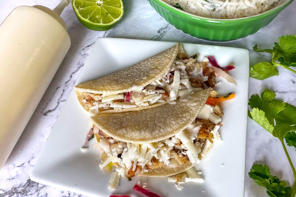 Two fish tacos topped with shredded cheese and creamy sauce on a square white plate, with lime, cilantro, and a bowl of rice in the background.