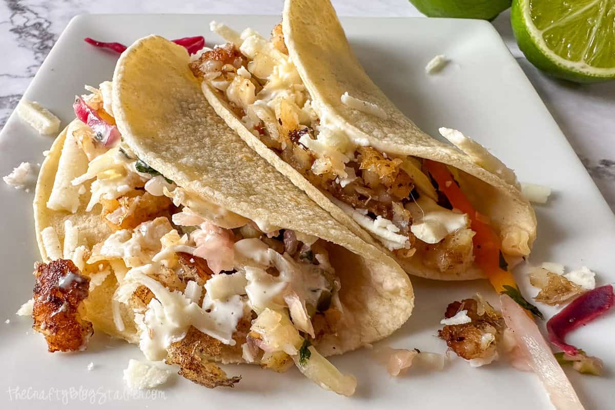 Two crispy fish tacos filled with shredded cabbage, cheese, and sauce on a white plate, with lime halves in the background.