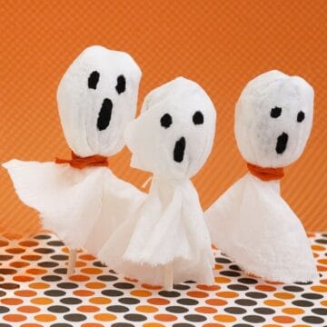 Three lollipops wrapped in white tissue with black ghost faces and orange string ties, displayed against an orange and polka dot background.