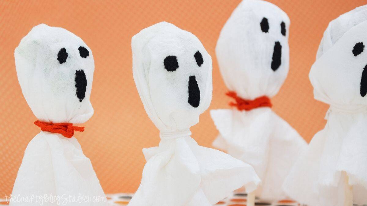 Four DIY ghost lollipop crafts made from tissues with black felt faces and orange string ties, displayed against an orange background.