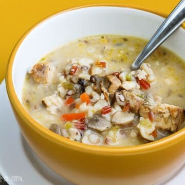 Bowl of creamy grilled chicken and wild rice soup with vegetables and a spoon, served in a yellow bowl on a white plate.