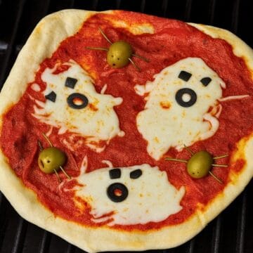 Halloween pizza topped with ghost-shaped melted cheese, black olive faces, and green olive spiders on tomato sauce.