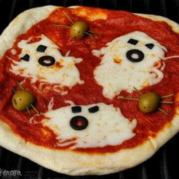 Halloween pizza topped with ghost-shaped melted cheese, black olive eyes and mouths, and green olive spiders on tomato sauce.