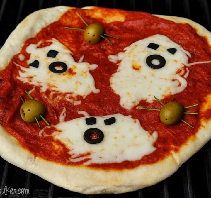 Halloween pizza topped with ghost-shaped melted cheese, black olive eyes and mouths, and green olive spiders on tomato sauce.