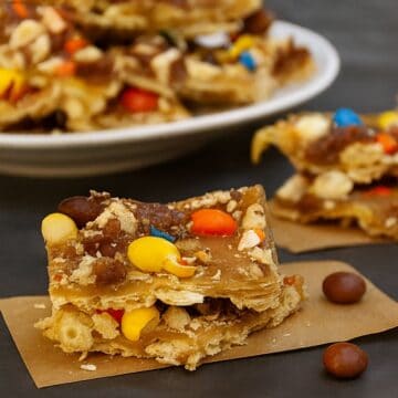 Close-up of Halloween candy saltine toffee topped with colorful M&M’s and caramel layers, stacked on parchment paper with more pieces in the background.