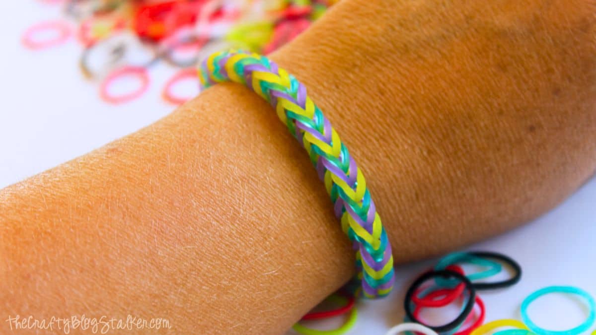 A handmade fishtail loom band bracelet in yellow, green, and purple rubber bands is worn on a wrist, with colorful loose bands scattered in the background.