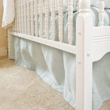 Close-up of a white crib with a light fabric skirt and mattress, showing the side detail on carpet in a nursery room.