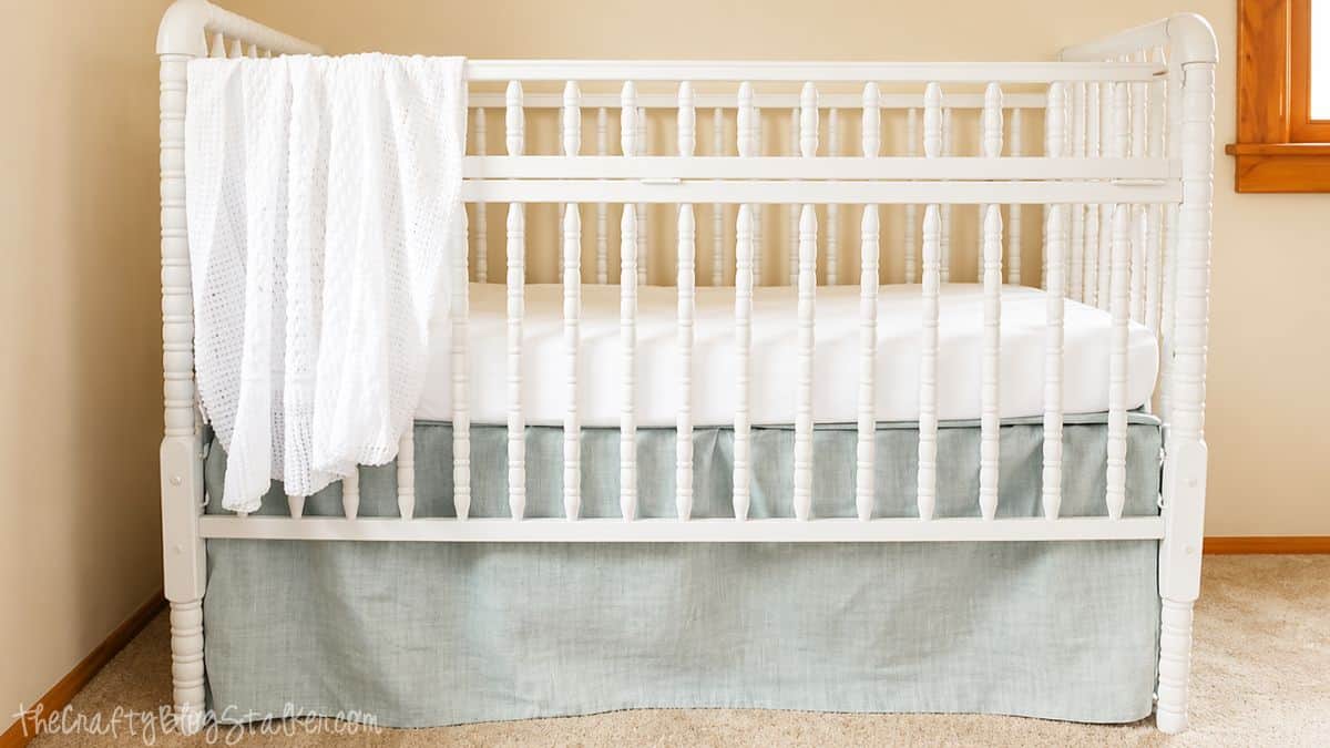 The Easiest DIY Crib Skirt Tutorial Ever! White crib with a light fabric skirt and mattress, with a soft blanket draped over the side in a nursery room.