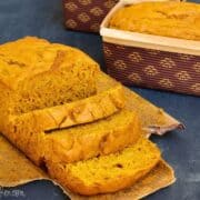 Loaf of homemade pumpkin bread with a few slices cut, sitting on parchment paper with another loaf in a brown patterned baking pan in the background.