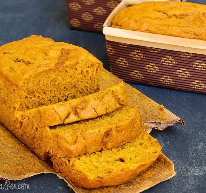 Loaf of homemade pumpkin bread with a few slices cut, sitting on parchment paper with another loaf in a brown patterned baking pan in the background.