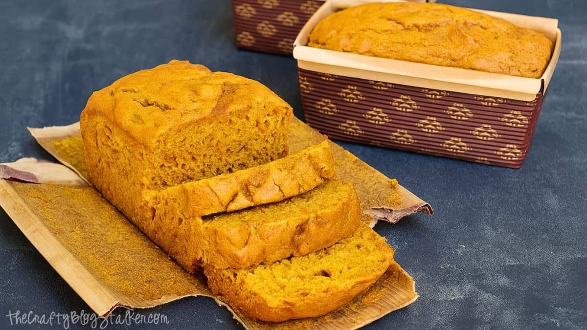 The Best ever Pumpkin Bread Recipe! Loaf of homemade pumpkin bread with a few slices cut, sitting on parchment paper with another loaf in a brown patterned baking pan in the background.