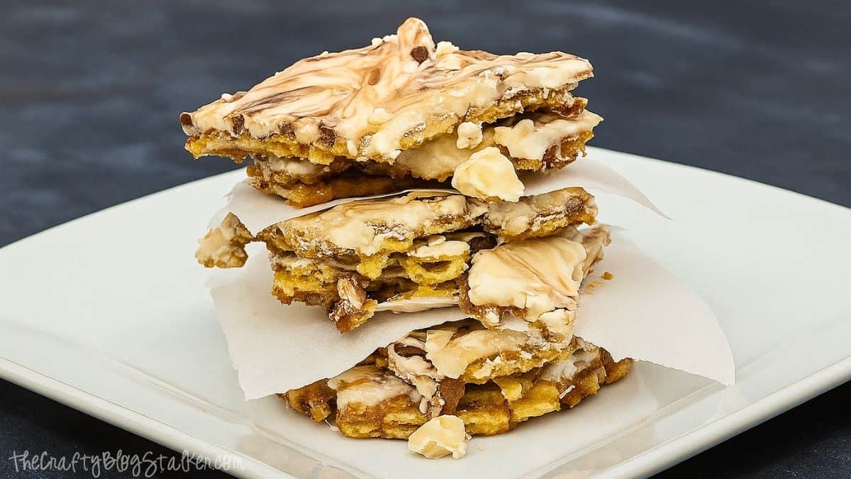 Stack of pumpkin pie toffee pieces layered with parchment paper on a white plate, showing a mix of toffee, nuts, and swirled white chocolate topping.