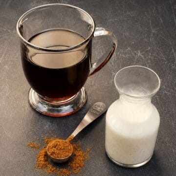 Glass mug of black coffee beside a small bottle of cream and a measuring spoon filled with pumpkin spice on a dark countertop.