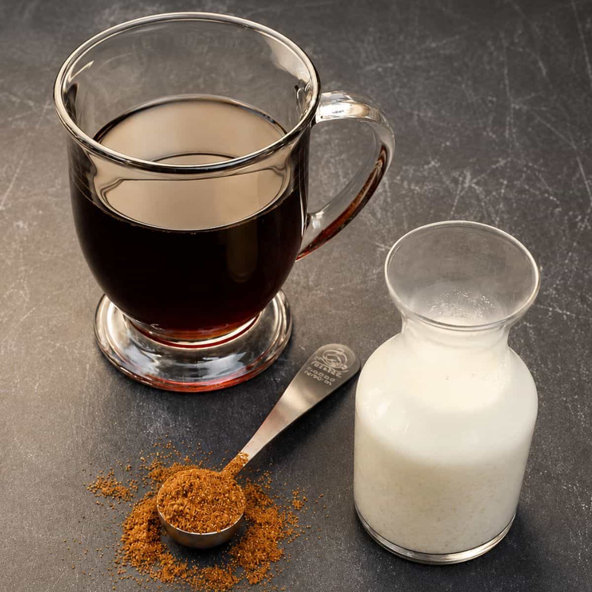 Glass mug of black coffee beside a small bottle of cream and a measuring spoon filled with pumpkin spice on a dark countertop.